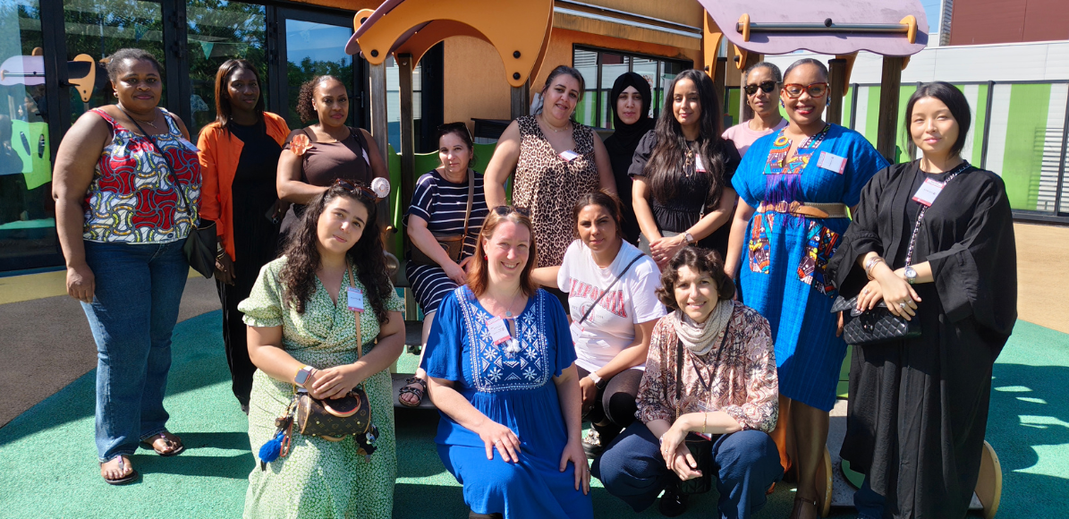 Group of diverse women posing for a photo on a sunny playground, with colorful play structures in the background.
