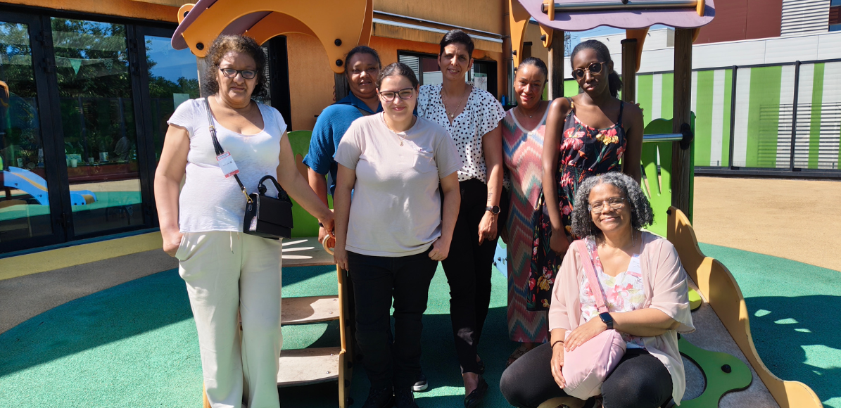 Seven women of diverse ages stand together on a colorful playground structure on a sunny day, posing for a group photo.