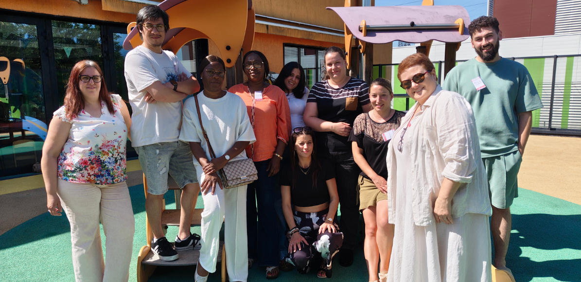 Group of diverse adults posing together on a playground outside a colorful building on a sunny day.