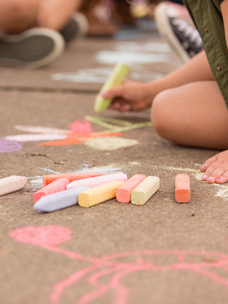 Child kneeling on concrete, drawing with sidewalk chalk; colorful chalk sticks scattered nearby and chalk shapes on the ground