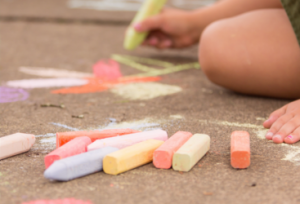 Child kneeling on a sidewalk and drawing with colorful chalk; chalk sticks are scattered nearby.