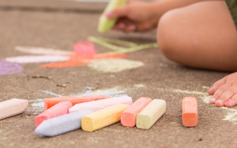 Child kneeling on concrete, drawing with sidewalk chalk; colorful chalk sticks scattered nearby and chalk shapes on the ground